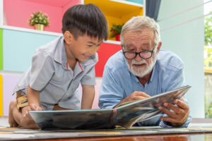 An older man and a little boy crouch side by side on the floor, smiling and reading a book together.