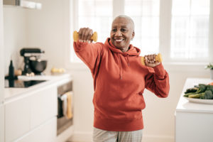 A woman holds hand weights while standing in the kitchen beside a plate of veggies, as she follows steps for improving health and wellbeing for seniors.