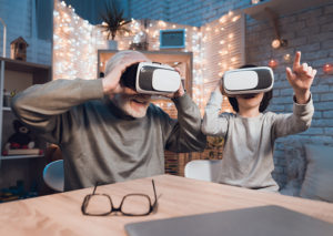 : A caregiver uses virtual reality in dementia care with an older man, as they both look into VR equipment and smile.