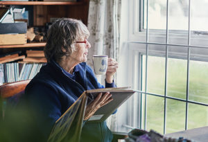 An older woman holds a photo album and a cup of coffee as she gazes out the window, experiencing some of the benefits linked to controlled clutter and dementia.