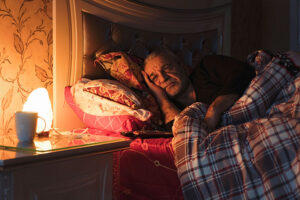 An older man in bed squints at the clock, struggling with changes in senior sleep patterns.