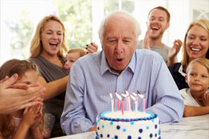 A family who worked together to come up with birthday party ideas for seniors gathers around an older man as he blows out the candles on his birthday cake.