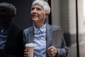 A woman balancing work and caregiving smiles confidently as she carries her laptop and a cup of coffee.
